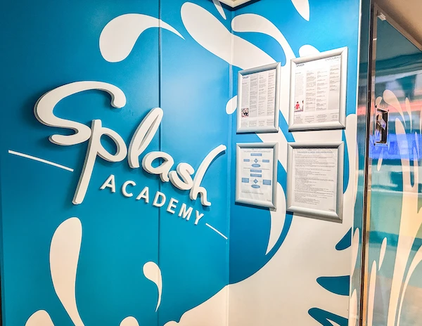 the entrance to the Splash Academy on a Norwegian Cruise Line ship, showcasing a vibrant blue wall decorated with white splash designs. The sign "Splash Academy" is prominently displayed along with framed documents, likely containing information about the programs and activities available for children at the academy.