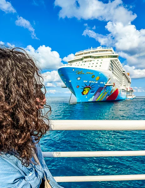 Kathy on a tender boat, looking towards a brightly decorated cruise ship, the Norwegian Breakaway, anchored near Great Stirrup Cay. The ship’s hull is vividly painted with colorful artwork featuring tropical motifs. The picture combines a clear blue sky and turquoise waters