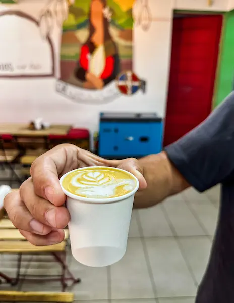 A hand holding a cup of coffee with latte art inside El Rincon Cafe in Puerto Plata on Umbrella Street.