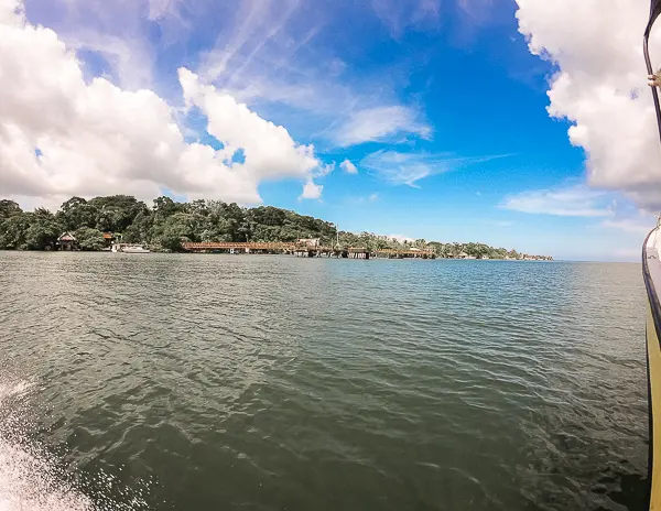 a view from a boat touring the Rio Dulce in Guatemala. The river is wide and calm, with a lush green shoreline and a blue sky with scattered clouds. The edge of the boat is visible on the right side of the image.