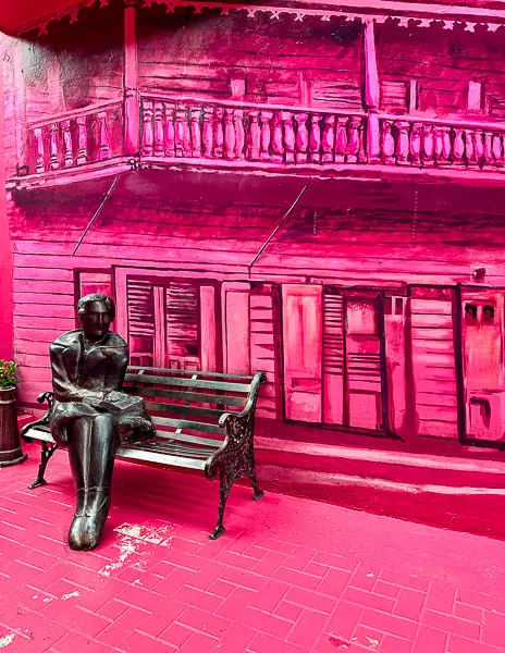 Statue of a woman sitting on a bench on Pink Street in Puerto Plata, with a pink mural in the background.