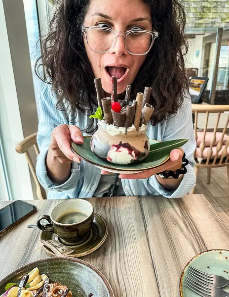 Woman (kathy) excitedly holding a decorative dessert with a cup of coffee on the table while dining on a cruise.

