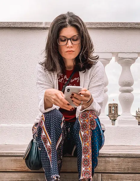 A woman (kathy) sits on a stone ledge outdoors, focused on her smartphone. She wears glasses, a casual white sweater, and colorful patterned pants, with a small bag by her side. In the background, architectural details, including white balusters and a distant building, can be seen.