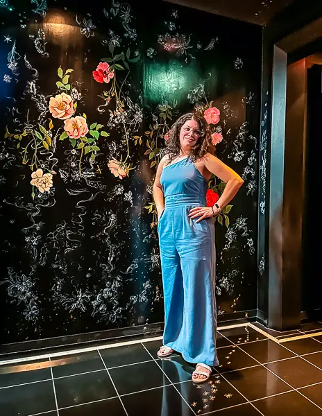 Woman (Kathy) in a blue jumpsuit posing against a floral wall, dressed for a chic night on a cruise.