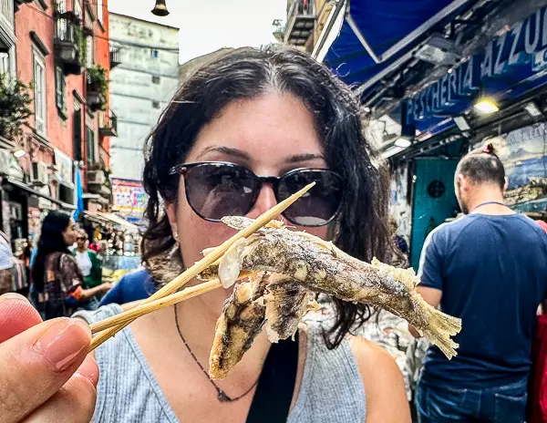 Kathy holding a small fish on a skewer while on a food tour in a bustling outdoor market, likely at a cruise port in Naples. She's wearing sunglasses and  is enjoying the local delicacy amidst a lively market scene in Naples Cruise Port on a food tour.