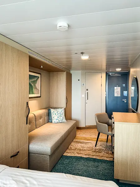 View of the interior of cabin 9644 on Utopia of the Seas, showing a sofa, desk, and modern wood-toned cabinets, with the door and entryway in the background.