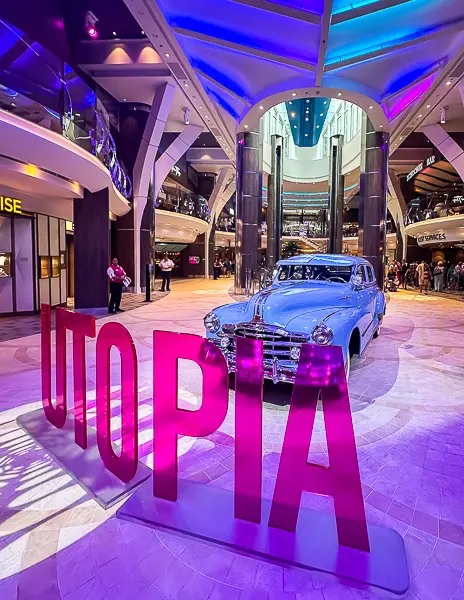 Promenade on Utopia of the Seas, featuring a classic blue car on display with vibrant pink 'UTOPIA' signage, surrounded by shops and illuminated with colorful lighting.