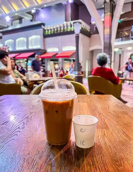 Iced coffee with a small cup of cream from Café Promenade on Utopia of the Seas, with seating in the background.