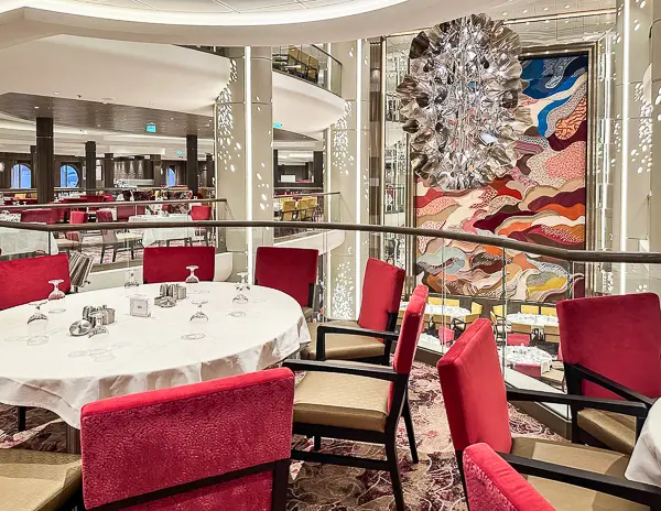 Main dining room on Utopia of the Seas, featuring elegant red chairs, a set dining table, and a stunning multi-story chandelier with intricate artwork in the background