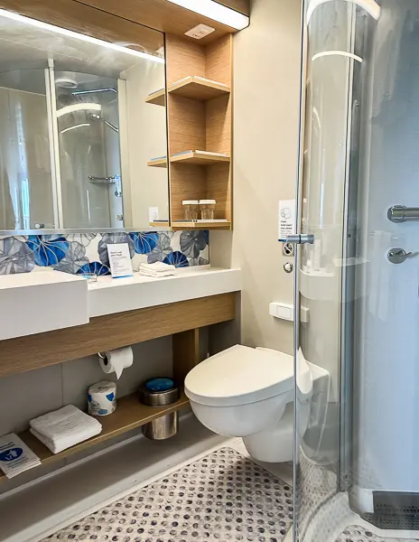 Bathroom in cabin 9644 on Utopia of the Seas, featuring a modern design with a glass-enclosed shower, vanity, and open shelving.