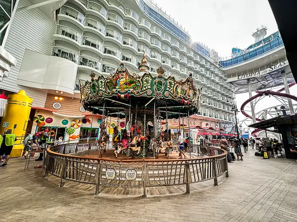 Carousel on The Boardwalk of Utopia of the Seas, surrounded by colorful attractions and stateroom balconies, with passengers enjoying the ride and walking nearby.