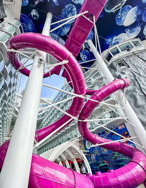 View of the Ultimate Abyss slide on Utopia of the Seas, featuring bright pink twisting tubes descending from the upper decks, set against the backdrop of stateroom balconies and a decorative mural.