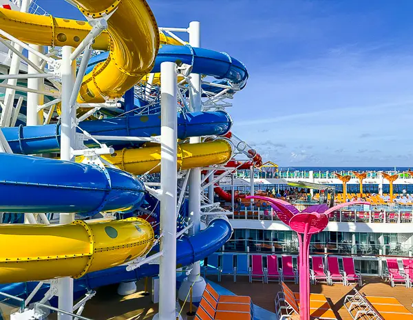 Bright blue, yellow, and red waterslides twisting down multiple decks on Utopia of the Seas, with a vibrant pool deck and ocean views in the background.