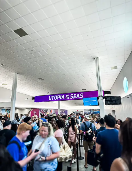 Crowd of passengers standing in line under a 'Utopia of the Seas' sign inside the cruise terminal at Port Canaveral, waiting to complete the boarding process.