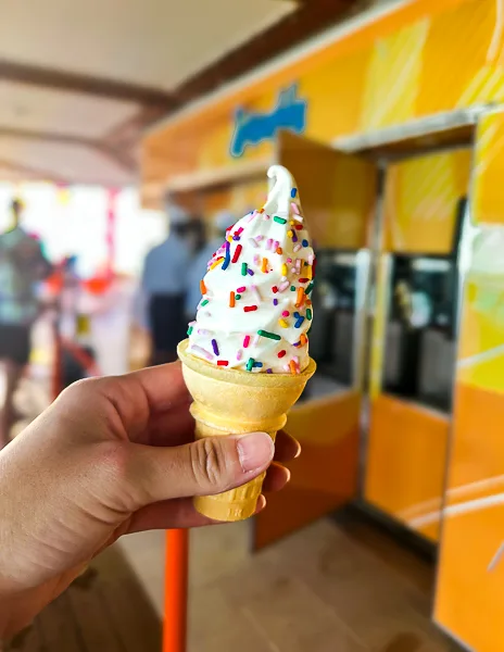 Soft-serve ice cream cone topped with rainbow sprinkles, held in front of the self-serve ice cream station on Utopia of the Seas.