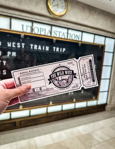 A hand holding train tickets for the 'Wild West Train Trip' experience, with Utopia Station’s schedule board in the background.