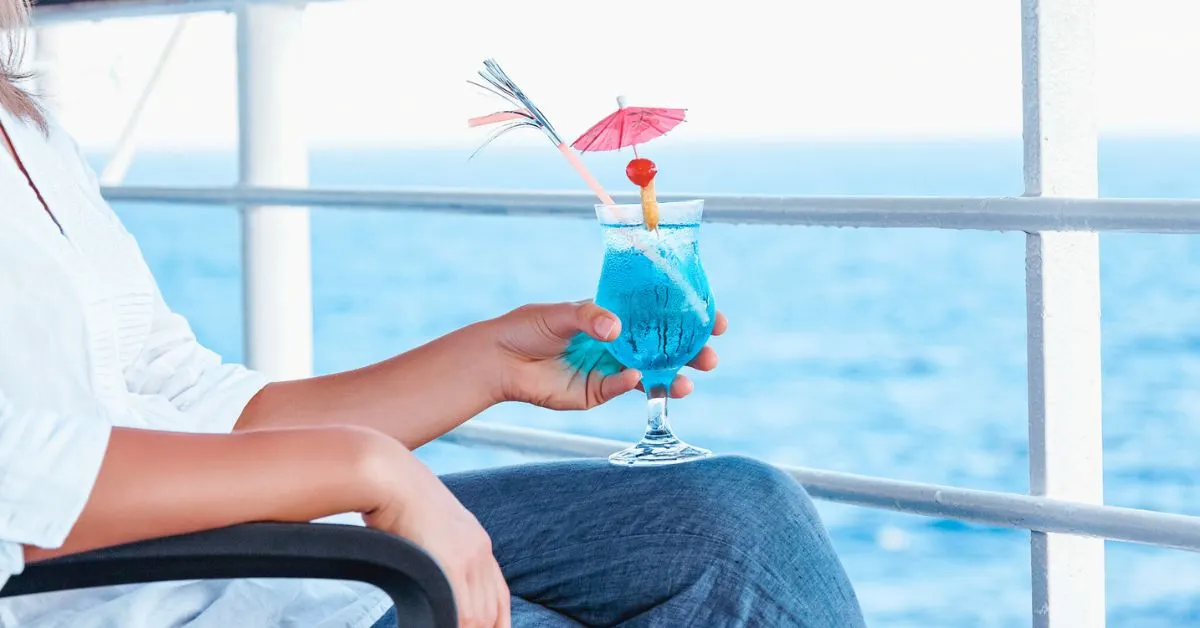 featured image: Close-up of a person holding a blue cocktail with a decorative umbrella and cherry, seated on a cruise ship balcony overlooking the ocean.