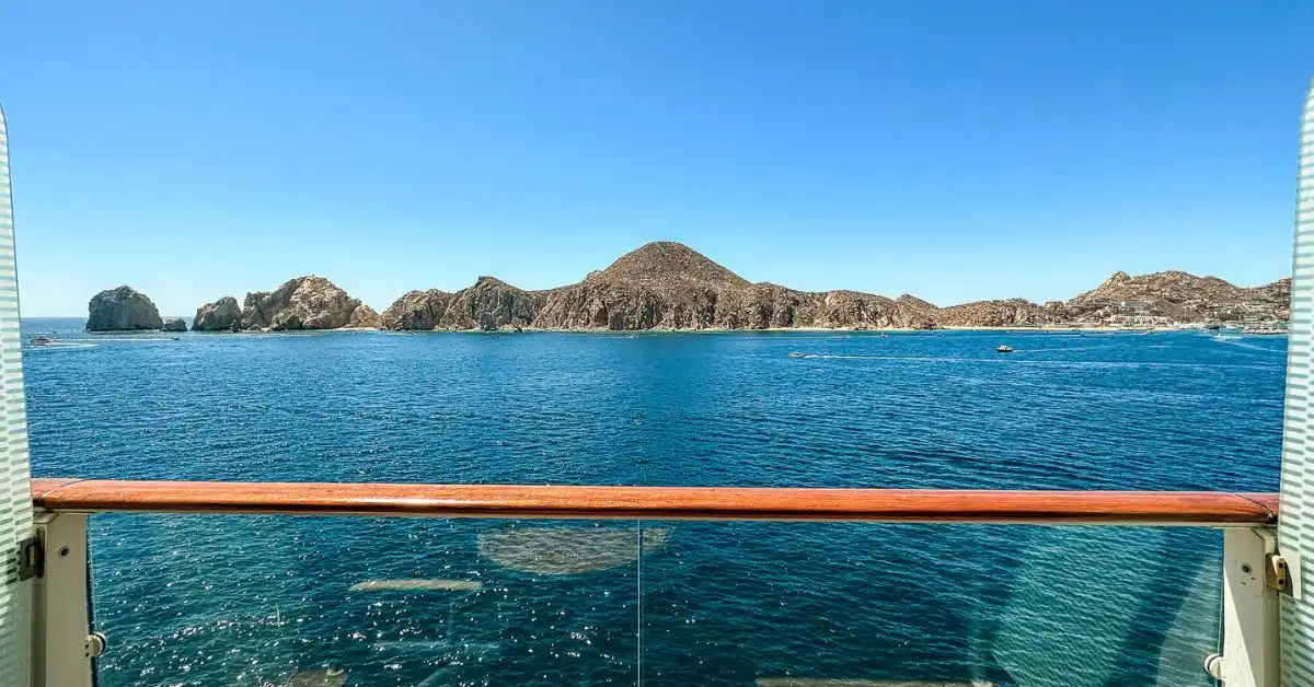 View of the Cabo San Lucas coastline from a cruise ship balcony, showcasing clear blue water and rocky cliffs under a bright sky.