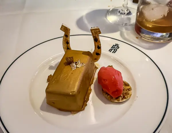 A gold bar-shaped dessert topped with edible gold and horseshoe-shaped chocolate, served with a side of pink sorbet on a white plate.