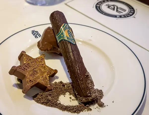 A chocolate dessert shaped like a cigar with a decorative band, served with chocolate dust and a chocolate sheriff’s badge on a white plate.