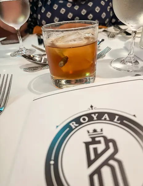 A glass of Sarsaparilla Old Fashioned cocktail served with a bullet embedded in the side of the glass, set on a white tablecloth with the Royal Railway menu in the background.