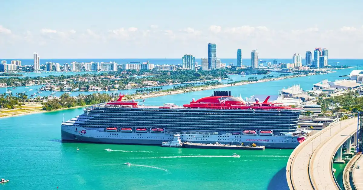 Virgin Voyages cruise ship docked in Miami with city skyline in the background, featuring the ship's sleek design and signature red accents.