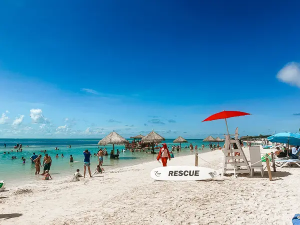 South Beach at CocoCay featuring calm turquoise water, beachgoers swimming, shaded in-water tables with umbrellas surrounding the floating bar, and a lifeguard station with a red umbrella.