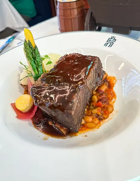 A plate of braised short ribs topped with rich brown sauce, served with beans, pickled vegetables, and asparagus on a white plate with the Royal Railway logo.