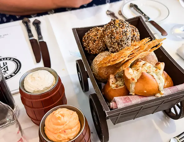 A bread basket served in a mini wagon, featuring a variety of breads including pretzels, seeded rolls, and crispy flatbread, accompanied by two small barrels of flavored butter on a white tablecloth.