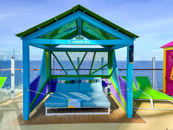 Vibrant blue and green casita labeled '12. Paradise Drive' on the deck of Royal Caribbean’s Utopia of the Seas, featuring cushioned seating and ocean views in the background.