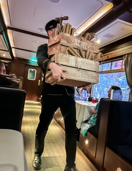 A conductor in period attire carrying a wooden crate labeled "ACME," adding to the immersive storytelling of the Royal Railway dinner experience.