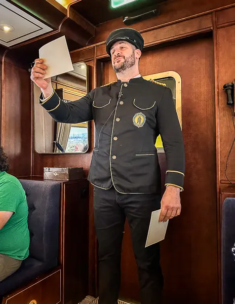 The conductor making an announcement during the Royal Railway experience, dressed in a formal uniform with gold trim, holding papers in one hand.