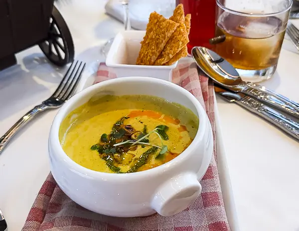 A white bowl of corn chowder garnished with herbs and oil, served with a side of crispy crackers on a red checkered napkin.