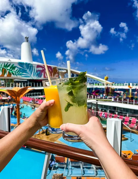 Two hands holding tropical drinks with paper straws on Royal Caribbean’s Utopia of the Seas, overlooking the colorful pool deck under a bright blue sky.