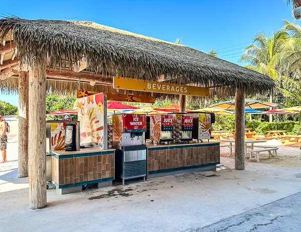 Beverage station at CocoCay with free ice water and juice dispensers under a thatched roof, surrounded by picnic tables and colorful umbrellas.