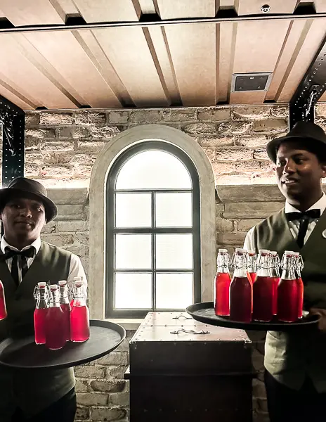 Two servers dressed in vintage Western attire holding trays of red "moonshine" drinks in glass bottles, standing in front of a rustic brick wall with an arched window.