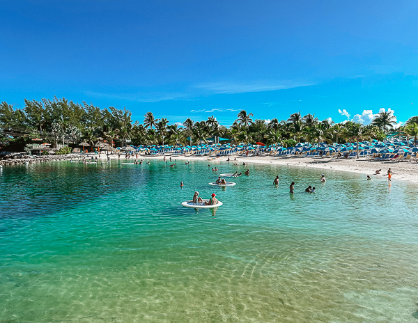 Harbor Beach at CocoCay with calm turquoise waters, beachgoers swimming, and round floating water mats scattered across the lagoon.