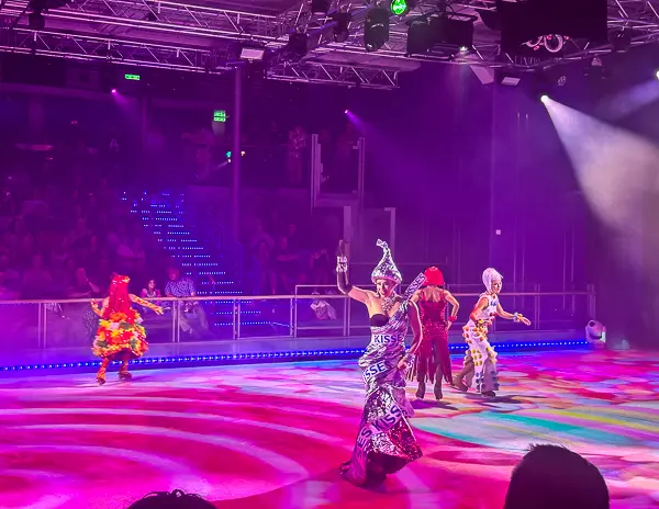 Performers in vibrant costumes skating during an ice show on a Royal Caribbean ship, illuminated by colorful lights and surrounded by an engaged audience.