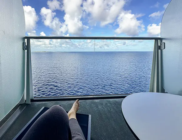 View from the Oceanview Balcony Cabin 9644 on Utopia of the Seas, showing clear blue skies, calm ocean water, and a passenger's legs resting on a reclining chair next to a small table.