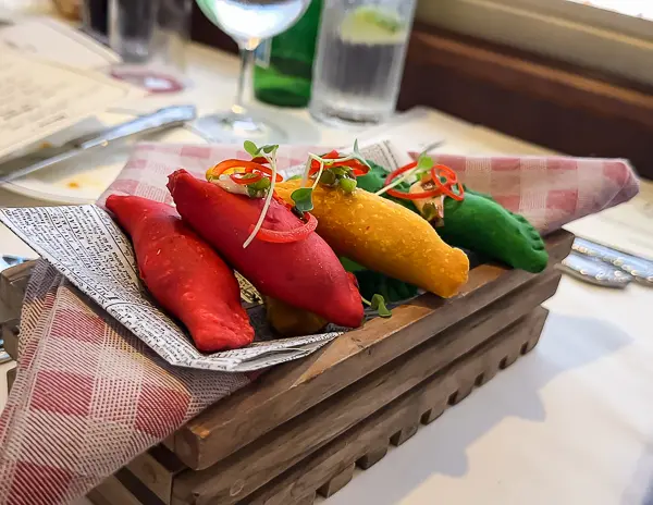 A trio of colorful empanadas in red, yellow, and green, served in a rustic wooden tray lined with faux newspaper and garnished with fresh herbs