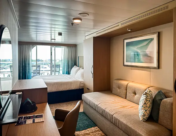 Interior view of Oceanview Balcony Cabin 9644 on Utopia of the Seas, featuring a bed, couch, desk, and balcony access with natural light streaming in.