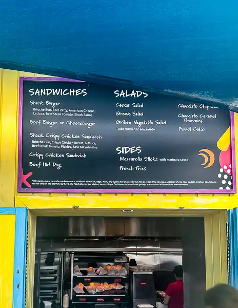 Snack Shack menu at CocoCay displaying sandwich options, salads, sides like mozzarella sticks and French fries, and desserts like brownies and funnel cake.