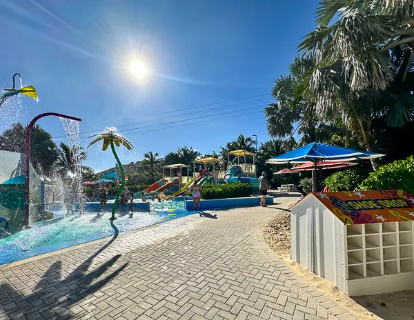 Side view of Splashaway Bay at CocoCay, showing interactive water play areas, slides, and seating shaded by umbrellas.