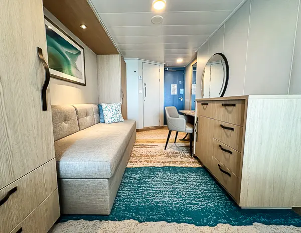 Interior view of Oceanview Balcony Cabin 9644 on Utopia of the Seas, featuring a sofa, dresser with a round mirror, and a desk with a chair leading to the cabin entrance.