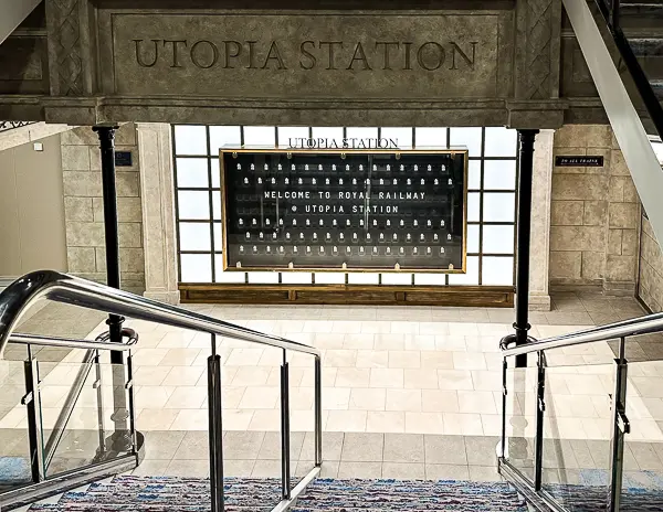 The entrance to Utopia Station on Utopia of the Seas, featuring a large welcome board with "Welcome to Royal Railway" displayed, visible from the staircase leading down to the station.