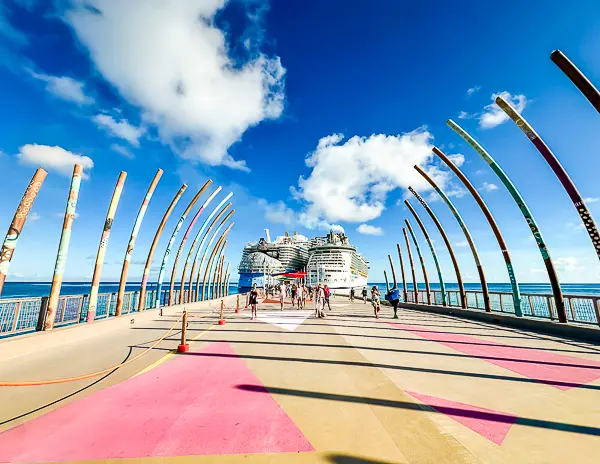 View of the pier at CocoCay with colorful archways, people walking toward a docked Royal Caribbean cruise ship under a bright blue sky.