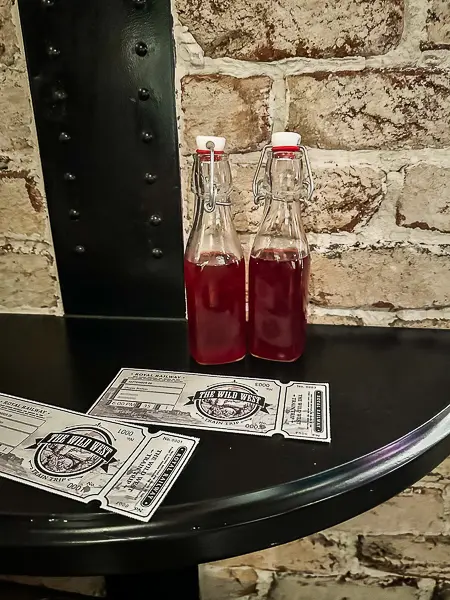 Two glass bottles of red "moonshine" drinks with flip-top lids, placed on a black table alongside vintage-style train tickets for the Wild West, set against a brick wall backdrop.