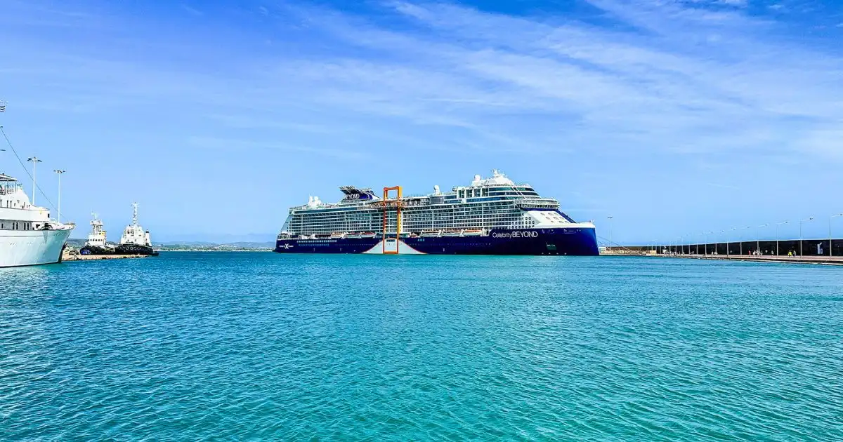 The Celebrity Beyond, an Edge-class cruise ship, docked in Katakolon, Greece. The ship features its signature Magic Carpet platform on the side, with calm turquoise waters surrounding it. Smaller vessels are nearby, and the sky is clear and blue.