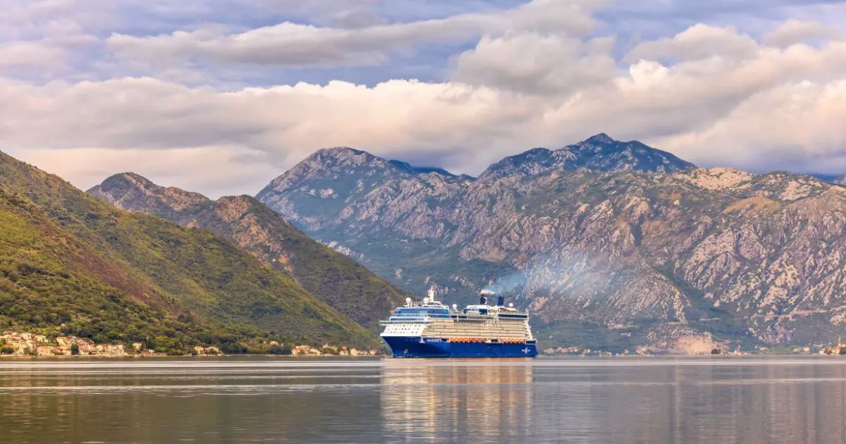 A Celebrity Cruises ship sails through a scenic fjord-like waterway, surrounded by towering mountains with lush greenery and rugged peaks under a cloudy sky.