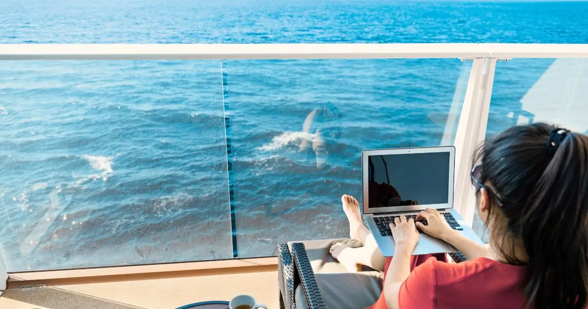 A person relaxing on a cruise ship balcony, working on a laptop while looking out at the ocean. The scene includes a clear glass railing, deep blue water, and a coffee cup nearby, suggesting a mix of leisure and productivity at sea.